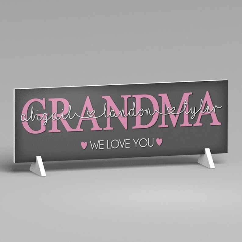 May include: A rectangular sign with the word "GRANDMA" in pink letters on a dark gray background. The names "abigail landon tyler" are above the word "GRANDMA". The phrase "WE LOVE YOU" is below the word "GRANDMA". The sign is supported by two white stands.