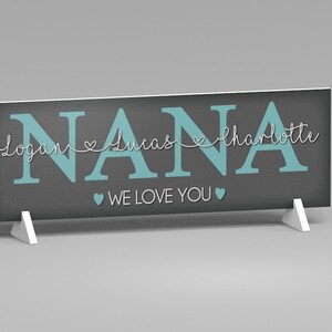 May include: A rectangular sign with the word "NANA" in teal block letters. Below the word "NANA" are the names "Logan", "Lucas", and "Charlotte" in a cursive font. The words "WE LOVE YOU" are below the names. The sign is gray and supported by two white stands.