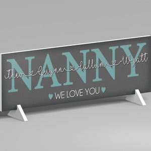 May include: A gray sign with the word "NANNY" in teal block letters. Below the word "NANNY" are the names "Theo", "Guynn", "Callum", and "Wyatt" in a white script font. The words "WE LOVE YOU" are below the names. The sign is supported by two white stands.