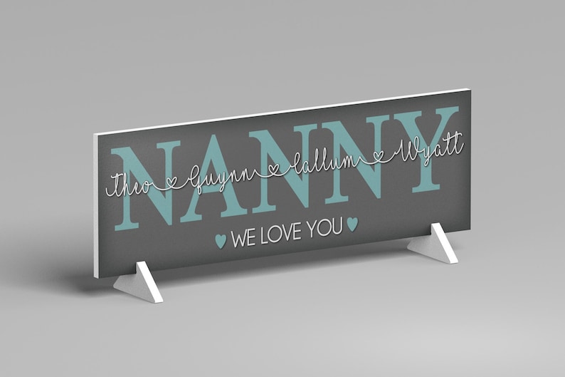 May include: A gray sign with the word "NANNY" in teal block letters. Below the word "NANNY" are the names "Theo", "Guynn", "Callum", and "Wyatt" in a white script font. The words "WE LOVE YOU" are below the names. The sign is supported by two white stands.