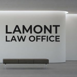 May include: A black and white sign with the text "LAMONT LAW OFFICE" on a white wall with a grey bench in front of it.