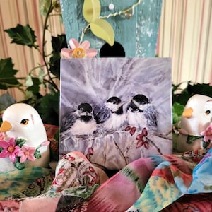May include: A watercolor painting of three black-capped chickadees perched on a branch with red berries. The painting is framed and displayed on a table with a colorful scarf and two white ceramic birds.