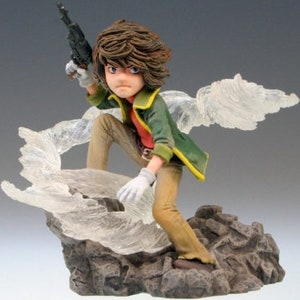 May include: A detailed figurine of a young man with brown hair, wearing a green jacket and brown pants, holding a gun and standing on a rocky base with a translucent effect.