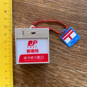 Peut inclure: Un porte-clés en plastique blanc avec un panneau de poste japonais rouge et blanc. Le panneau indique "JP Post" et "ゆうゆう窓口" qui se traduit par "Guichet de service postal".