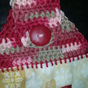 Peut inclure: Un gros plan sur un bouton rouge cousu sur une serviette de cuisine en crochet avec du fil rose, rouge et marron. La serviette a un fond blanc avec un motif de flocons de neige et de bouteilles de vin.