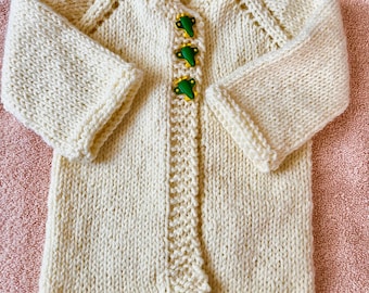 May include: A cream-colored hand-knitted cardigan featuring long sleeves, a textured knit pattern, and three green airplane-shaped buttons down the front. The garment has a ribbed hem and cuffs with a decorative open-stitch detail along the button placket.
