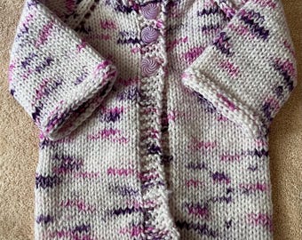 May include: A hand-knitted long-sleeve cardigan made from white yarn with variegated flecks of purple and pink. The garment features a round neckline and a front button closure with three round, light purple buttons that have a spiral pattern.