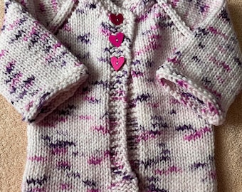 May include: A hand-knitted long-sleeve cardigan featuring a variegated yarn pattern in shades of white, pink, and dark purple. The garment includes three heart-shaped buttons in bright pink, arranged vertically along the center front opening.