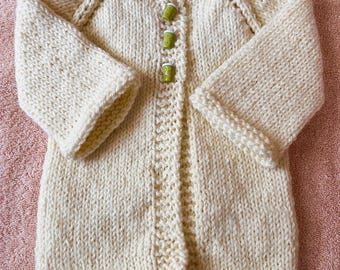 May include: A cream-colored hand-knitted cardigan featuring long sleeves, a V-neckline, and a front button closure. The garment is secured with three small, cylindrical green buttons with white tops, and it displays a textured knit pattern.