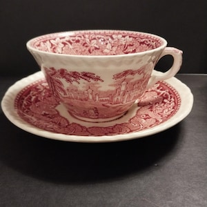 May include: A cream-colored teacup and saucer set featuring a detailed maroon transferware pattern. The design depicts an architectural landscape with buildings and trees, complemented by a floral border on the rim and interior of the cup.
