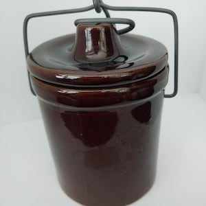 May include: A brown ceramic crock with a lid and a wire handle. The crock has a glossy finish and a cylindrical shape. The lid has a small knob in the center. The wire handle is attached to the sides of the crock.