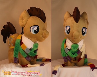 Può includere: Un cavallo di peluche marrone e beige che indossa una sciarpa a righe verde, viola e bianca. Il cavallo ha una criniera e una coda marroni e occhi blu.