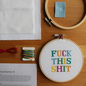 May include: A cross-stitch embroidery kit with a white fabric hoop, red embroidery floss, a small spool of multi-coloured embroidery floss, a needle, and a piece of blue felt. The hoop contains a white fabric with the phrase "FUCK THIS SHIT" stitched in colourful thread.