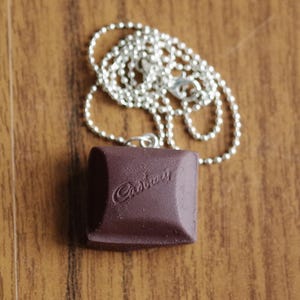 Può includere: Una collana a catena a sfera color argento con un ciondolo marrone scuro. Il ciondolo è di forma quadrata con bordi arrotondati e la scritta "Cadbury" è in rilievo sul davanti. La collana è appoggiata su una superficie di legno.