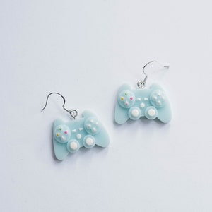 Op de afbeelding: Een paar lichtblauwe hangende oorbellen in de vorm van een videogamecontroller. De controller heeft witte knoppen en een kleurrijk ontwerp.