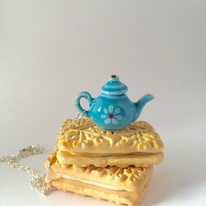 May include: A miniature blue teapot with a white and red floral design sits atop a stack of three cream-colored, square biscuits. A silver chain necklace is draped near the biscuits. The teapot has gold accents.