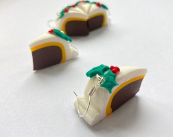 May include: A pair of dangle earrings featuring a white, yellow, and brown cake slice with a green holly leaf and red berries on top. The earrings are made of polymer clay and have silver hooks.