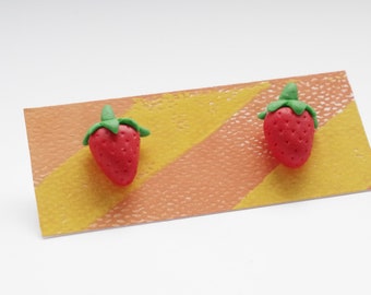 May include: A pair of red and green clay strawberry earrings on a striped orange and yellow background.