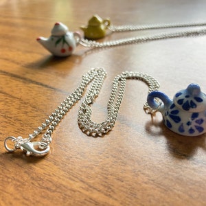 Può includere: Una collana a catena d'argento con tre ciondoli a forma di teiera. Una teiera è bianca con accenti rossi, una è verde e una è blu e bianca.