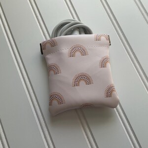 May include: A small, light pink pouch with a repeating pattern of brown and gold sun designs. The pouch is open at the top, revealing a coiled gray cable. The background is a white surface with vertical lines.