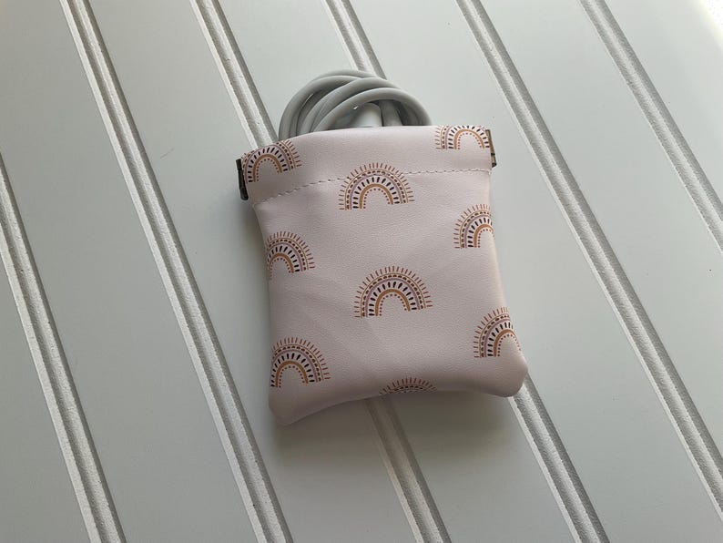 May include: A small, light pink pouch with a repeating pattern of brown and gold sun designs. The pouch is open at the top, revealing a coiled gray cable. The background is a white surface with vertical lines.