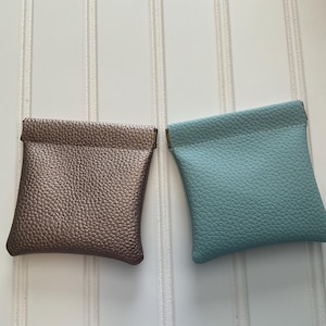 May include: Two small, square pouches. One is a metallic bronze color, and the other is a light teal. Both have a textured, pebbled surface and a simple, folded top. The pouches are likely for small items like coins or jewelry.