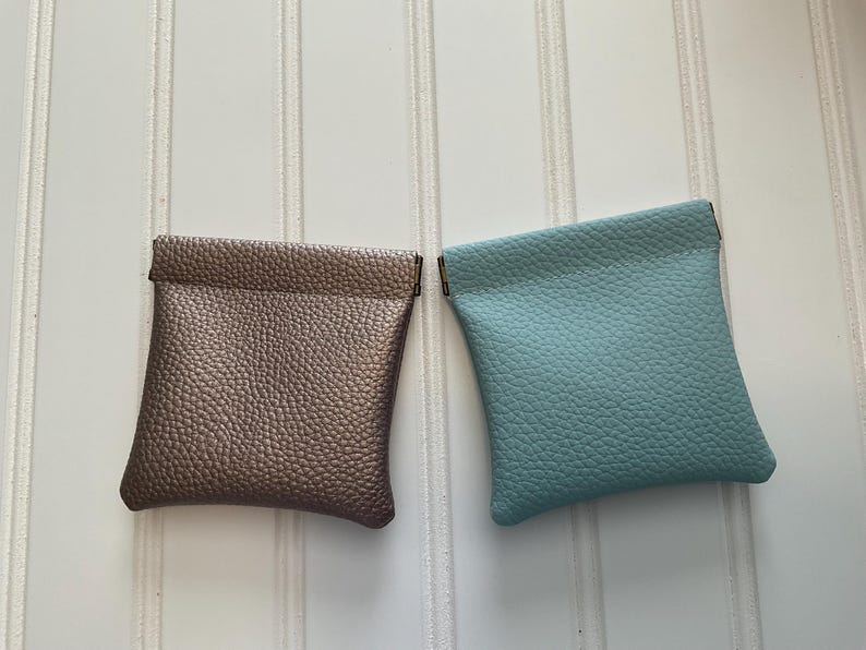May include: Two small, square pouches. One is a metallic bronze color, and the other is a light teal. Both have a textured, pebbled surface and a simple, folded top. The pouches are likely for small items like coins or jewelry.