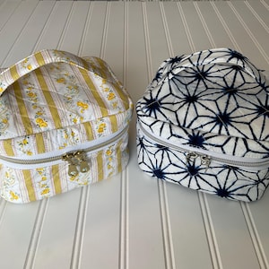 May include: Two quilted cosmetic bags. One is yellow and white with a floral and stripe pattern, and the other is white with a navy blue geometric design. Both have a zipper closure and a top handle.
