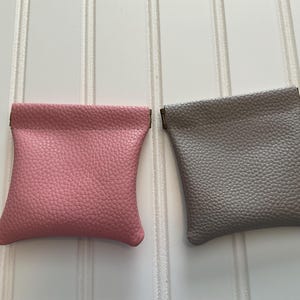 May include: Two small, textured leather pouches. One is a dusty rose color, and the other is a light gray. Both pouches have a rectangular shape with a curved bottom and a folded top. The pouches are displayed on a white, vertical-slatted surface.