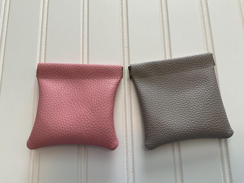 May include: Two small, textured leather pouches. One is a dusty rose color, and the other is a light gray. Both pouches have a rectangular shape with a curved bottom and a folded top. The pouches are displayed on a white, vertical-slatted surface.