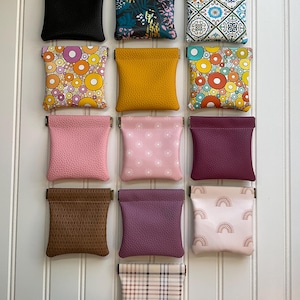 May include: A collection of small, square coin purses in various colors and patterns. The purses include solid black, floral, geometric, and patterned designs. The purses are made of leather and fabric.