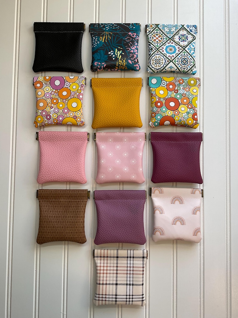 May include: A collection of small, square coin purses in various colors and patterns. The purses include solid black, floral, geometric, and patterned designs. The purses are made of leather and fabric.