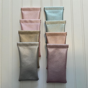 May include: Eight rectangular pouches with a textured, metallic finish arranged in two vertical columns. The colors include shades of pink, gold, silver, and light blue. Each pouch features a simple, flat design with a folded top edge.
