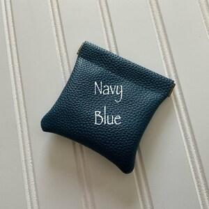 May include: A navy blue leather pouch with a textured surface. The text "Navy Blue" is written in white script across the front of the pouch. The pouch is square-shaped and has a gold-colored trim.