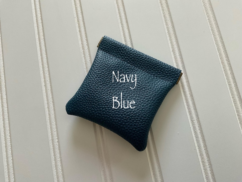 May include: A navy blue leather pouch with a textured surface. The text "Navy Blue" is written in white script across the front of the pouch. The pouch is square-shaped and has a gold-colored trim.