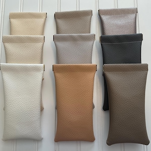 May include: A collection of nine leather eyeglass cases in various neutral colors, including cream, tan, gray, and black. Each case has a textured surface and a rectangular shape with a fold-over top.