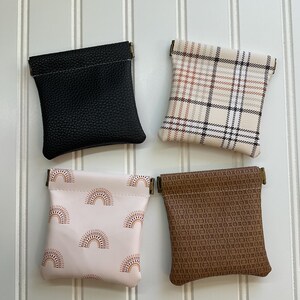 May include: Four small pouches in a square arrangement. The pouches are black, plaid, pink with rainbow pattern, and brown with a geometric pattern. Each pouch has a metal clasp at the top. The pouches are displayed against a white, vertical-slatted background.