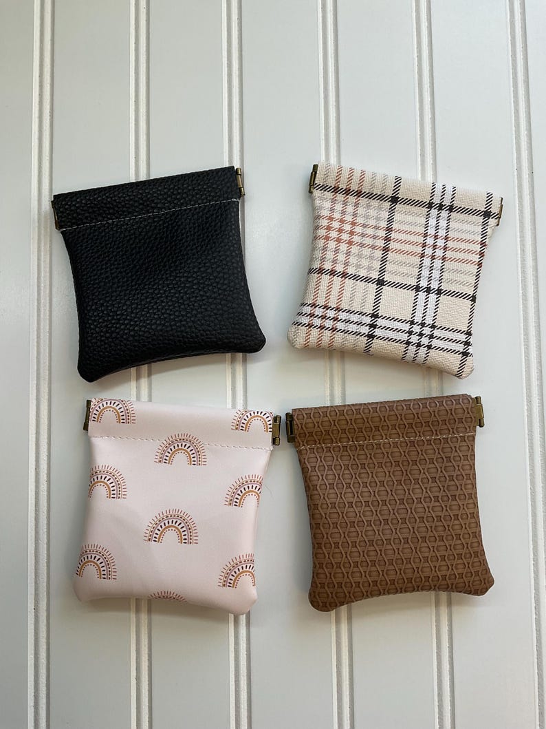 May include: Four small pouches in a square arrangement. The pouches are black, plaid, pink with rainbow pattern, and brown with a geometric pattern. Each pouch has a metal clasp at the top. The pouches are displayed against a white, vertical-slatted background.