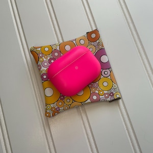 May include: A bright pink wireless earbud case rests on a patterned fabric pouch. The pouch features a colorful design of overlapping circles in yellow, orange, and purple. The items are on a white, slatted surface.