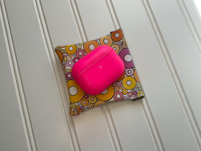 May include: A bright pink wireless earbud case rests on a patterned fabric pouch. The pouch features a colorful design of overlapping circles in yellow, orange, and purple. The items are on a white, slatted surface.