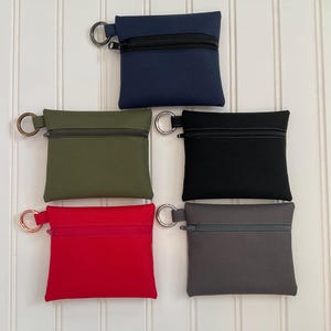 May include: Five square zippered pouches in navy blue, olive green, black, red, and gray. Each pouch features a top zipper closure and a metal D-ring attached to the side for hanging or attachment, providing a compact storage solution.