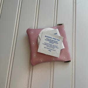 May include: A small, pink, floral-patterned pouch with a metal clasp, holding a white alcohol prep pad packet. The packet has blue text that reads "ALCOHOL PREP PAD" and other details. The pouch is on a white, paneled surface.