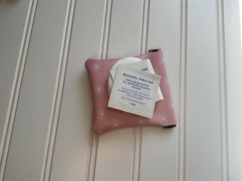 May include: A small, pink, floral-patterned pouch with a metal clasp, holding a white alcohol prep pad packet. The packet has blue text that reads "ALCOHOL PREP PAD" and other details. The pouch is on a white, paneled surface.
