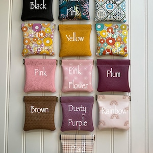 May include: A collection of small, square pouches in various colors and patterns. Each pouch is labeled with a color name, including black, teal/pink, blue, yellow, pink, plum, brown, dusty purple, and rainbow. The pouches are displayed against a white background.