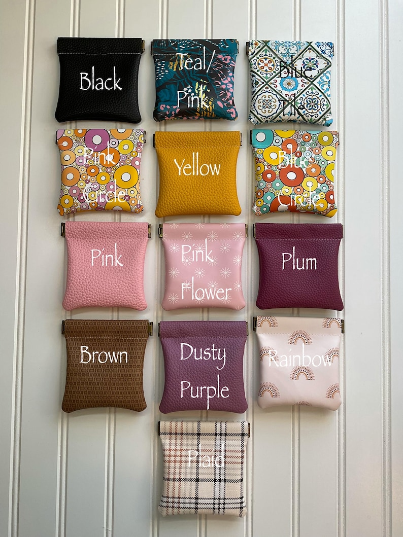 May include: A collection of small, square pouches in various colors and patterns. Each pouch is labeled with a color name, including black, teal/pink, blue, yellow, pink, plum, brown, dusty purple, and rainbow. The pouches are displayed against a white background.