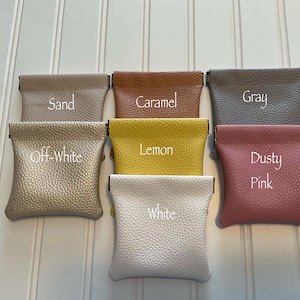 May include: A collection of small, textured pouches in various colors, including sand, caramel, gray, off-white, lemon, dusty pink, and white. Each pouch has a rectangular shape with a fold-over top and is labeled with its color name.
