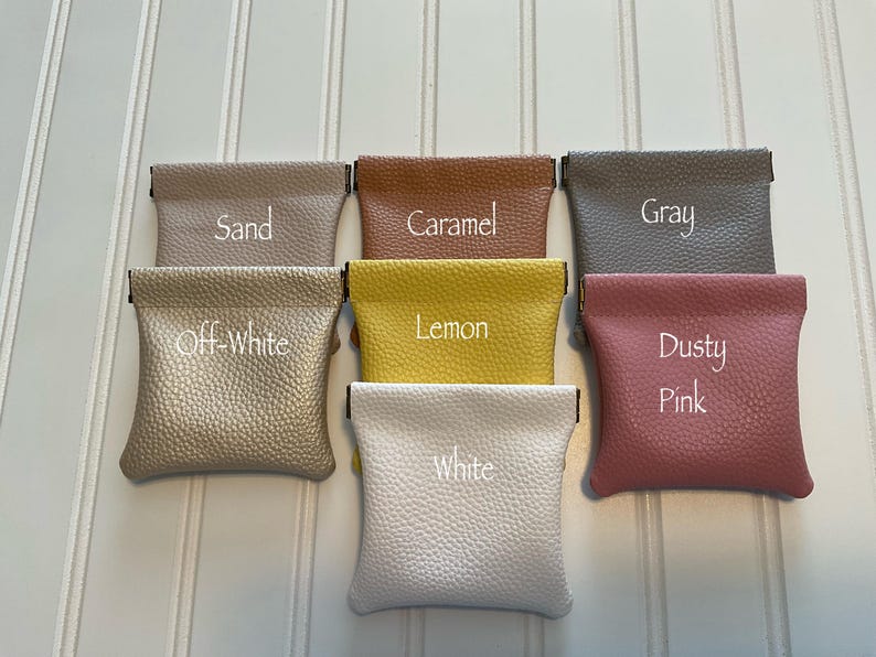 May include: A collection of small, textured pouches in various colors, including sand, caramel, gray, off-white, lemon, dusty pink, and white. Each pouch has a rectangular shape with a fold-over top and is labeled with its color name.