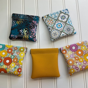 May include: Five small, square pouches in various patterns and colors. Designs include a tropical leaf print, a geometric tile pattern, and colorful circles. One pouch is solid mustard yellow.