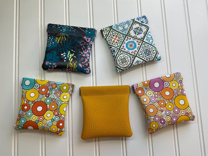 May include: Five small, square pouches in various patterns and colors. Designs include a tropical leaf print, a geometric tile pattern, and colorful circles. One pouch is solid mustard yellow.