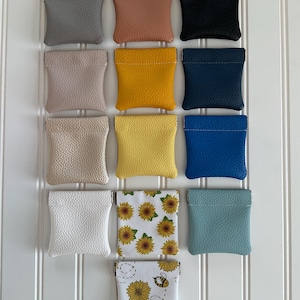 May include: A collection of thirteen small, square squeeze pouches arranged in a grid. The pouches feature various solid colors including gray, tan, black, mustard, navy, beige, yellow, blue, white, and teal, plus two white pouches with sunflower and bee prints.